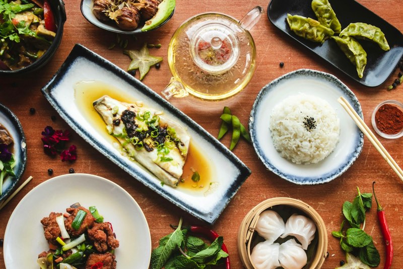 Table filled with Chinese dishes: steamed fish, dumplings, rice, dim sum and crispy chicken